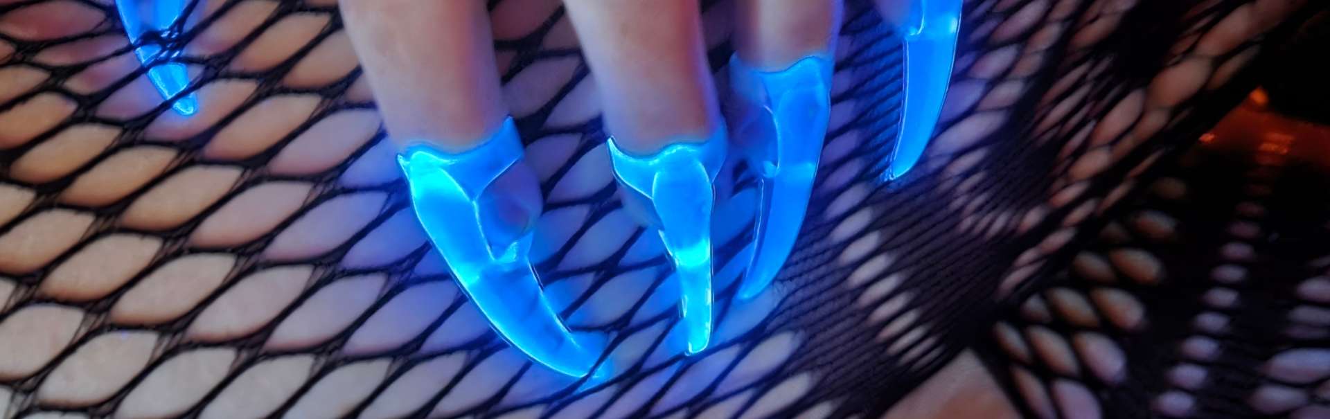 Glowing Crystal Fetish Claws lit up by UV, being worn n adult hand which is resting on leg in fishnet stockings.