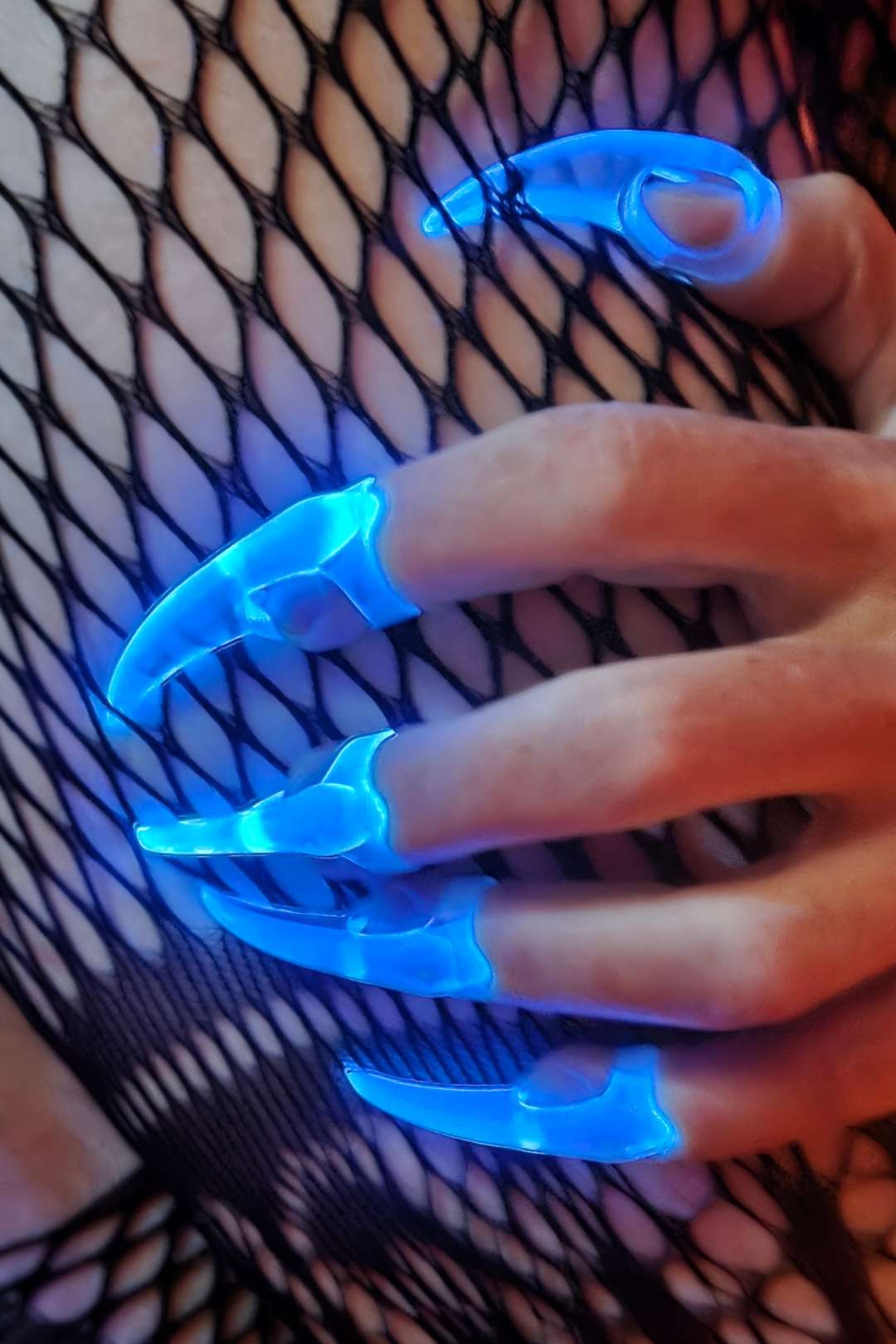 Glowing Crystal Fetish Claws lit up by UV, being worn n adult hand which is resting on leg in fishnet stockings.