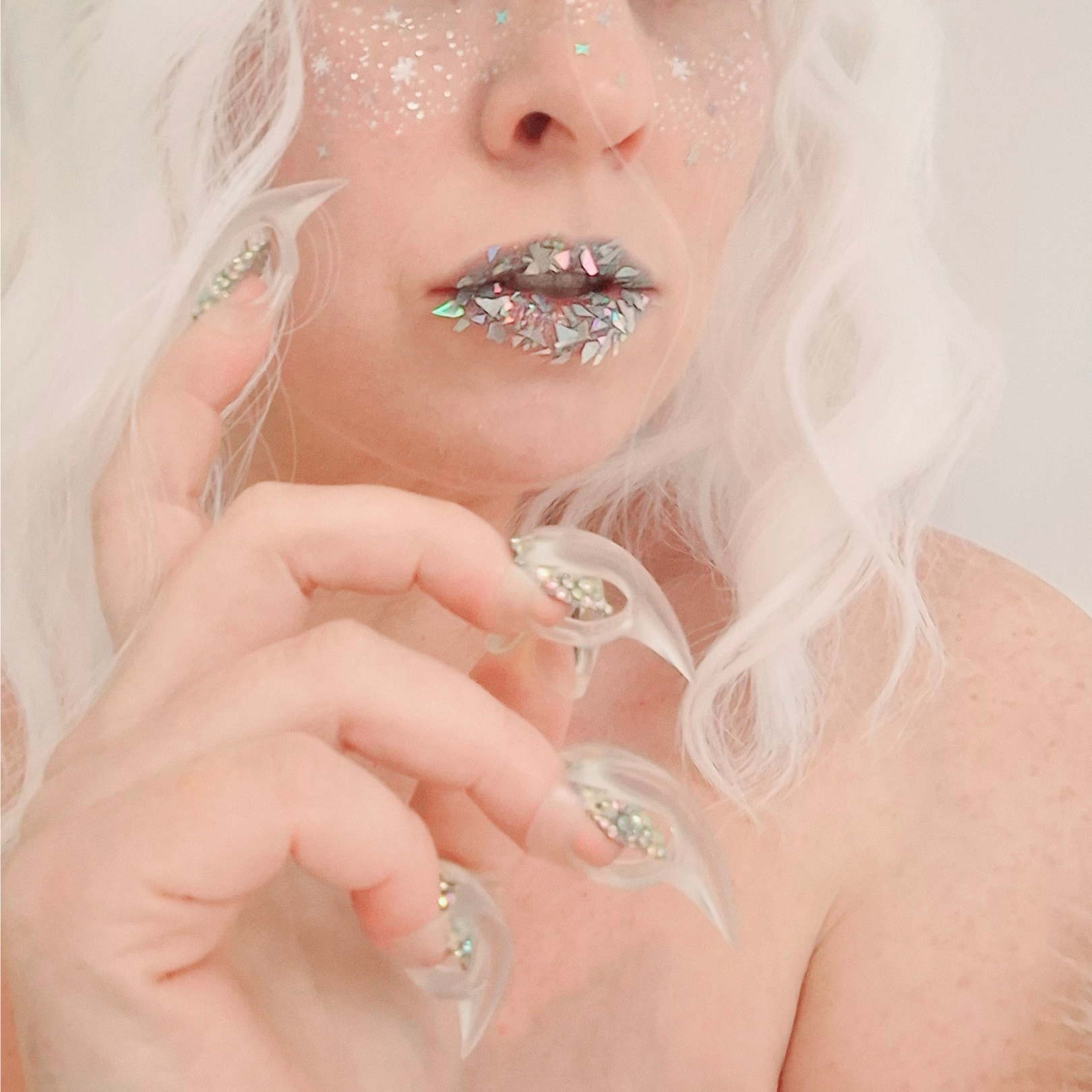 Person with glittery makeup and white hair wearing clear adult fingertip claws resembling talons.