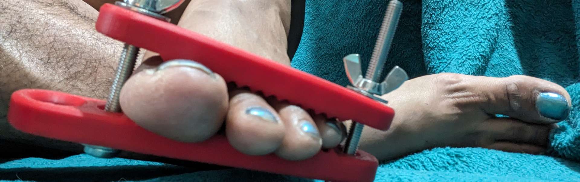 Red Flesh Clamp holding mainicured bare toes. Model is wearing no clothes and sitting on a blue towel.